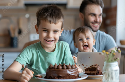 A young boy in green shirt eating chocolate cake, while his father and baby brother sit at the table watching him from behind.