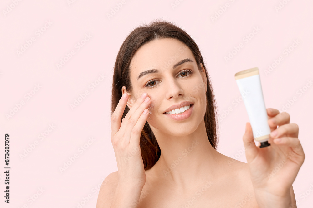 Beautiful young woman with tube of sunscreen applying cream on face against pink background Beautiful young woman with tube of sunscreen applying cream on face against pink background