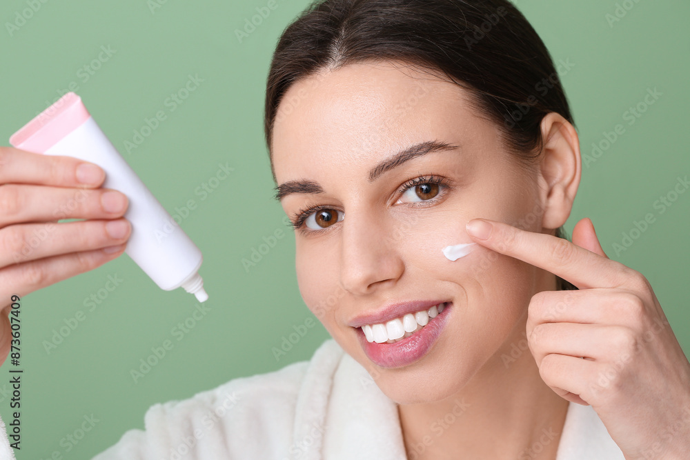 Beautiful young woman in robe applying face cream on green background Beautiful young woman in robe applying face cream on green background