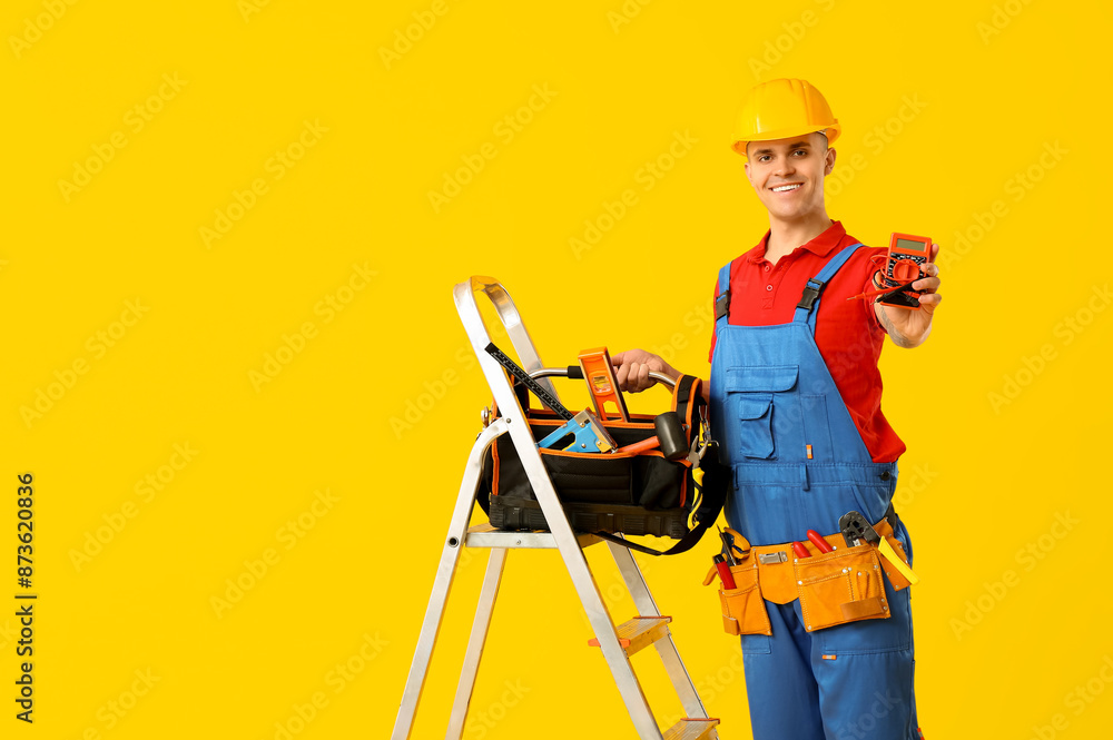 Young male electrician with multimeter and tool bag standing on stepladder against yellow background Young male electrician with multimeter and tool bag standing on stepladder against yellow background