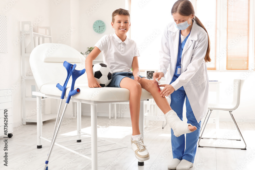 Obraz premium Smiling little boy with wounded knee and soccer ball visiting doctor in clinic