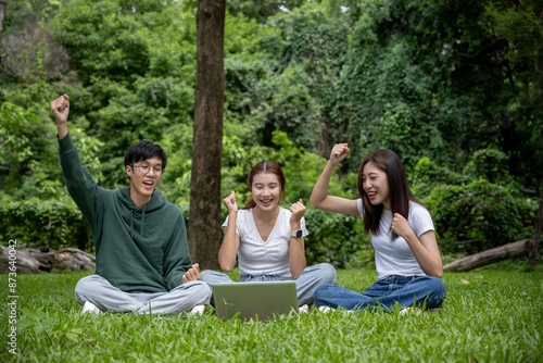 A group of cheerful young Asian college students is checking their exam scores on a laptop and celebrating together in a park, sitting on the grass, raising their hands and showing their fists.