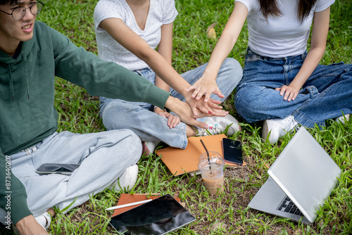 A close-up image of a group of cheerful Asian college students is putting their hands together, cheering up, and showing teamwork while sitting on the grass in the green park.