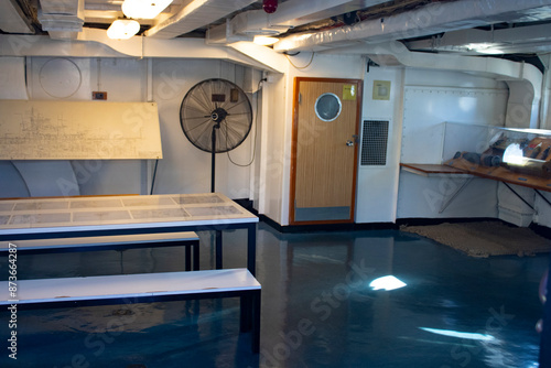 Captivating interior of an Australian naval warship room with white table, benches, and
 metallic floor. Subtle dim yellow light adds a unique ambiance against the backdrop of 
Sydney sea.