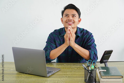 Smiling young asian man sit work at wooden desk with pc laptop. Achievement business career lifestyle concept. hold hands folded in prayer