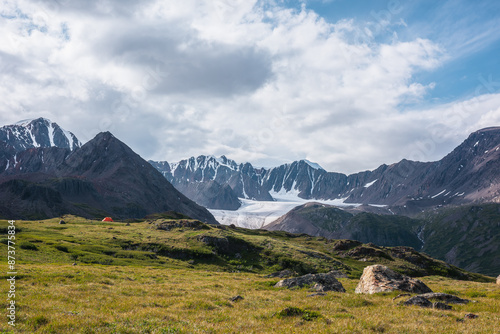 Dramatic scenery with orange tent in wide alpine valley among green hills and rocks with view to big glacier tongue and large snow-capped mountain range under clouds in blue sky in changeable weather.