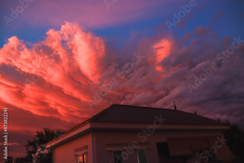 A dramatic summer sunset sky with orange and pink clouds over a house, creating a peaceful yet striking atmosphere © MarieXMartin