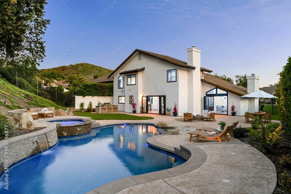 Fototapeta premium luxury house with a swimming pool and patio area, surrounded by greenery and a clear evening sky.