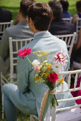 A man in a light blue suit sits on a white chair adorned with a colorful floral arrangement during an outdoor wedding ceremony. photo