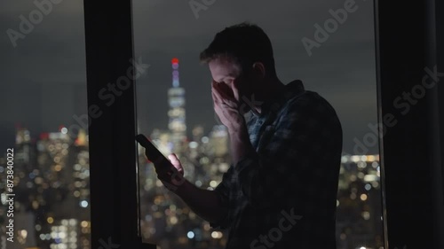 Man or businessman holding smartphone reading and looking at screen at night with metropolitan city buildings in the background. Unhappy and disappointed or feeling bad. Texting and shaking his head