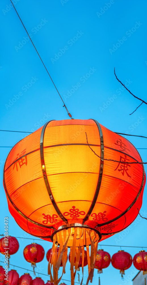 Obraz premium Close-up detail of an internally lit Chinese New Year lantern hanging on the street in London's Chinatown in Soho, with other lanterns in the background below, under a clear blue twilight sky.