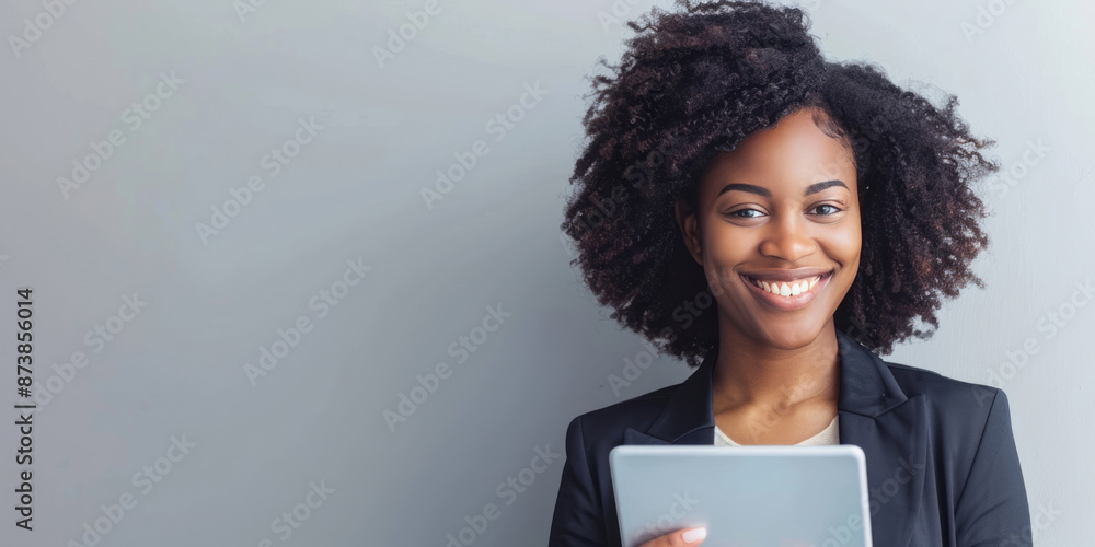 A curly-haired businesswoman, professionally dressed and holding a tablet, smiles against a light grey backdrop. A curly-haired businesswoman, professionally dressed and holding a tablet, smiles against a light grey backdrop.