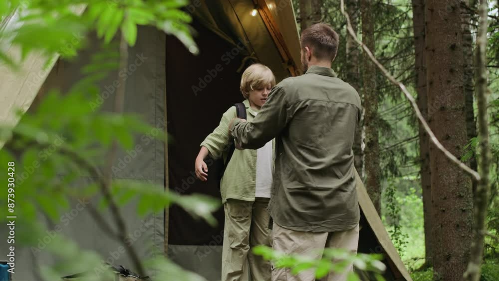 Loving Caucasian father putting backpack on his sons shoulders while preparing to go trekking together, standing beside cozy camping tent with yellow light bulbs in lush forest Loving Caucasian father putting backpack on his sons shoulders while preparing to go trekking together, standing beside cozy camping tent with yellow light bulbs in lush forest
