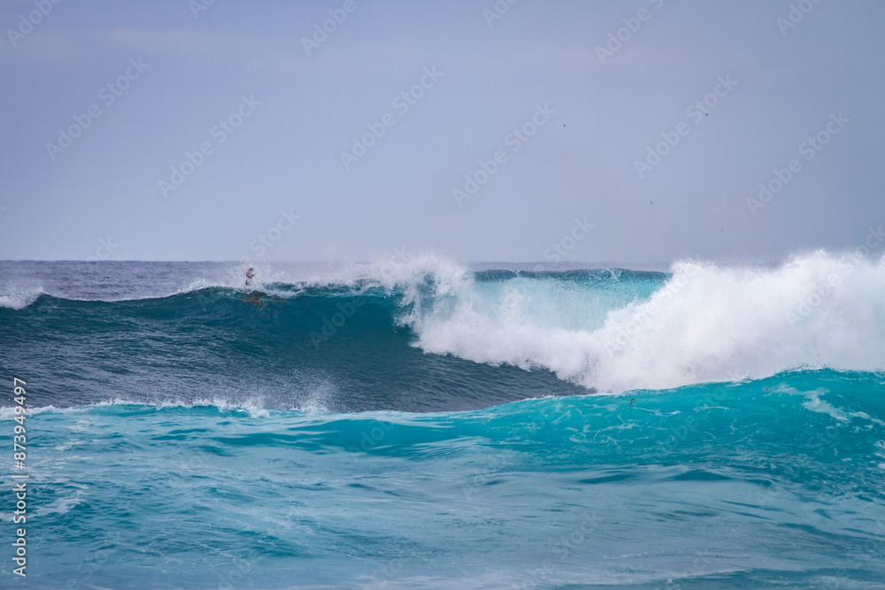 Waves coming ashore on a sunny day and surfers ready to surf the waves. Wall of blue wave with white foam coming ashore where surfers are surfing. Atlantic Ocean. Tenerife, Canary Islands, Spain. Waves coming ashore on a sunny day and surfers ready to surf the waves. Wall of blue wave with white foam coming ashore where surfers are surfing. Atlantic Ocean. Tenerife, Canary Islands, Spain.