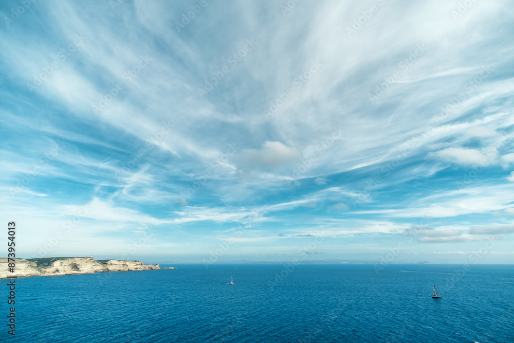 Yachts sailing past the limestone cliffs in the bay of Bonifacio on the south coast of Corsica with the coastline of Sardinia in the distance under dramatic summer sky Yachts sailing past the limestone cliffs in the bay of Bonifacio on the south coast of Corsica with the coastline of Sardinia in the distance under dramatic summer sky