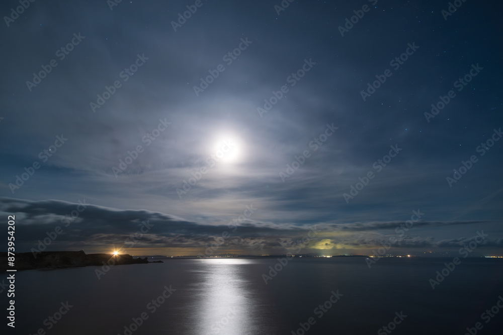 Fototapeta premium The full moon lights up the night sky over the cliffs of Bonifacio and Mediterranean sea on the south coast of Corsica with the lights from the coastline of Sardinia in the distance
