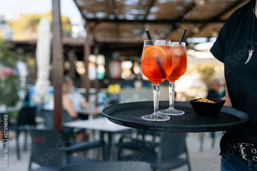 Waiter holding tray with two spritz cocktails and snacks. Waiter is carrying a tray with two spritz cocktails and a bowl of snacks in a busy outdoor restaurant