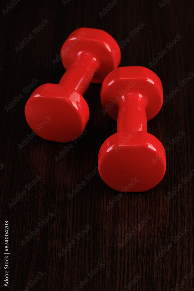 Two shiny red dumbbells lie on a dark wooden surface. Selective focus. Vertical orientation. Two shiny red dumbbells lie on a dark wooden surface. Selective focus. Vertical orientation.