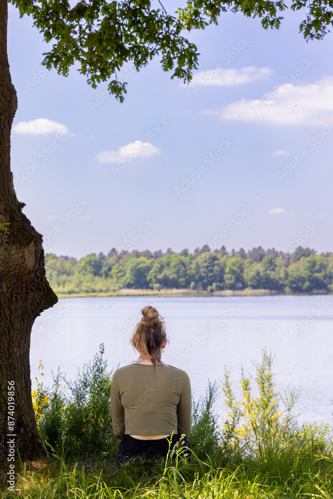Fototapeta premium Woman sits by the lake, enjoying serene nature and tranquility, under a tree in a peaceful outdoor setting.