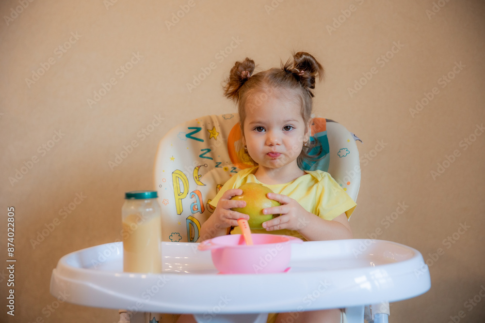 A little girl is sitting on a high chair and eating a green apple at home life style A little girl is sitting on a high chair and eating a green apple at home life style