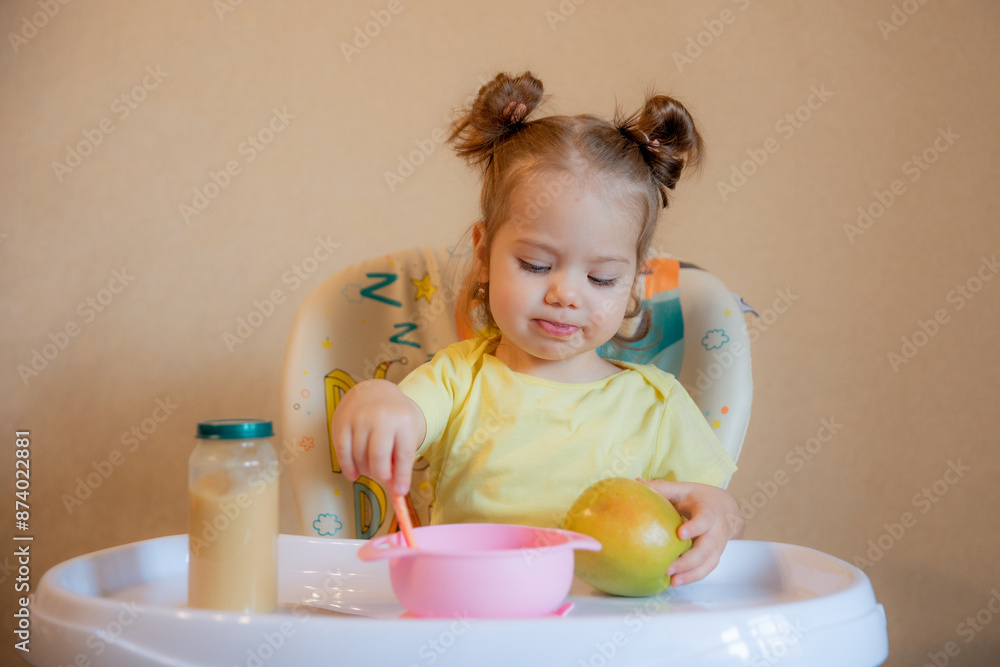 A little girl is sitting on a high chair and eating mashed fruit at home A little girl is sitting on a high chair and eating mashed fruit at home