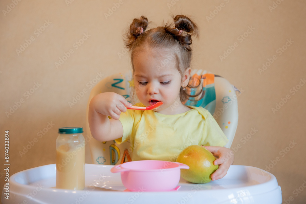 A little girl is sitting on a high chair and eating mashed fruit at home A little girl is sitting on a high chair and eating mashed fruit at home