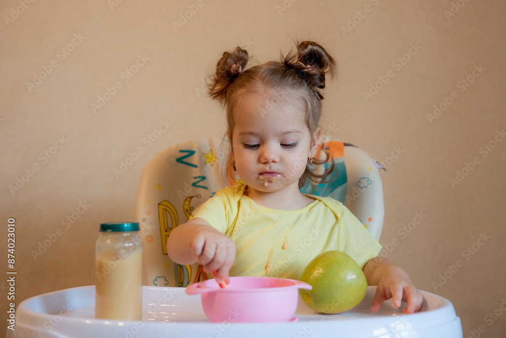 A little girl is sitting on a high chair and eating mashed fruit at home A little girl is sitting on a high chair and eating mashed fruit at home