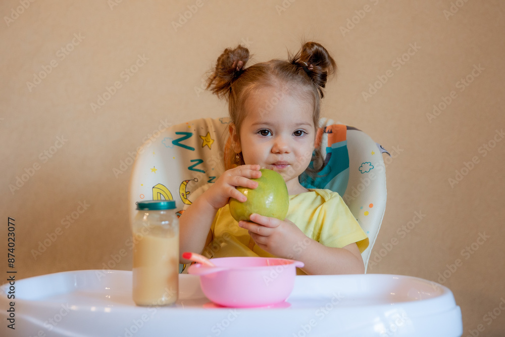 A little girl is sitting on a high chair and eating a green apple A little girl is sitting on a high chair and eating a green apple