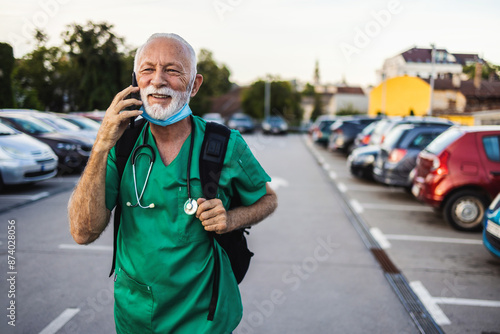 Professional male Caucasian nurse in scrubs talking on a mobile phone, while on his way to work. Male doctor consulting patient online while searching for his car in the parking lot after work.