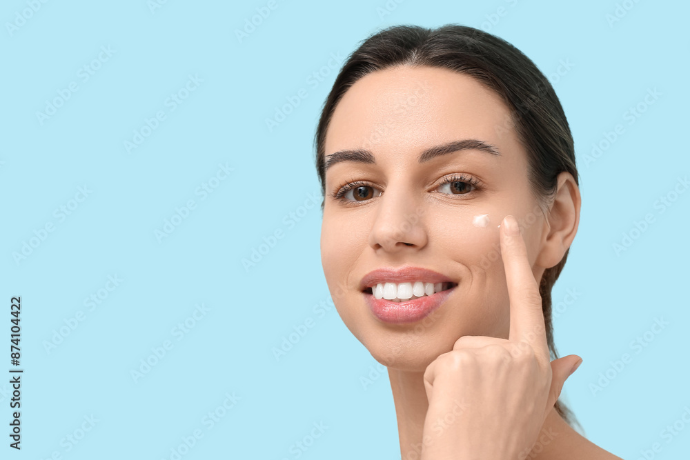 Beautiful young woman applying face cream against blue background Beautiful young woman applying face cream against blue background