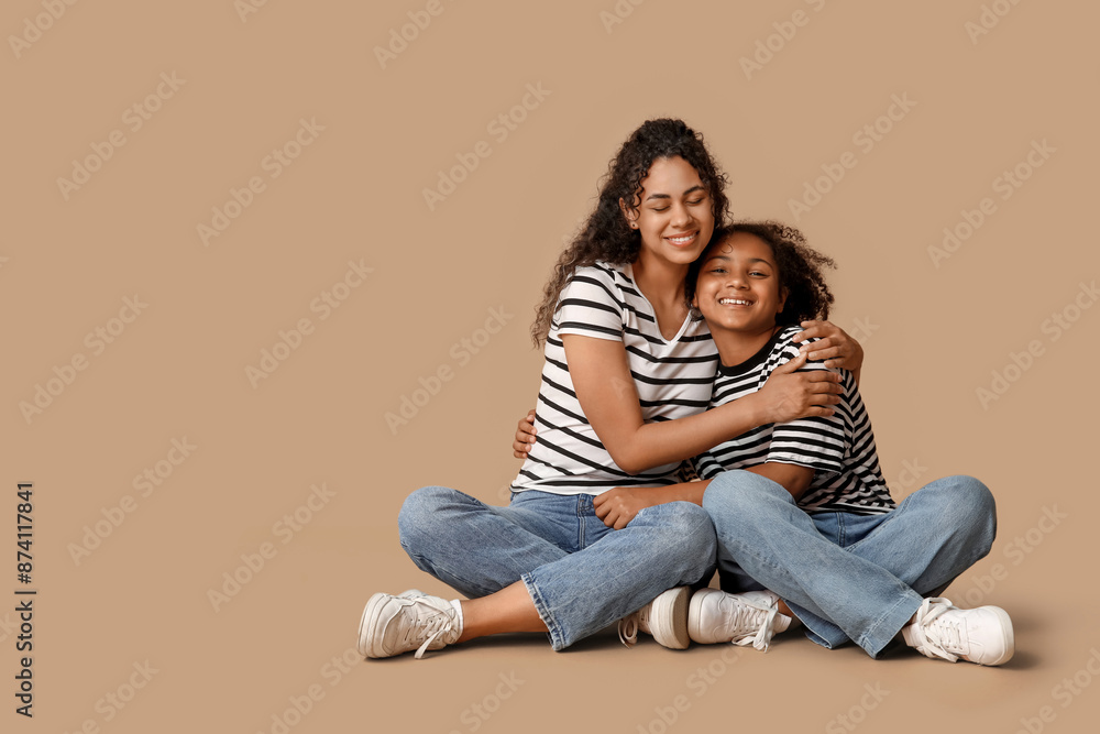 Beautiful young happy African-American mother with her daughter sitting on beige background Beautiful young happy African-American mother with her daughter sitting on beige background