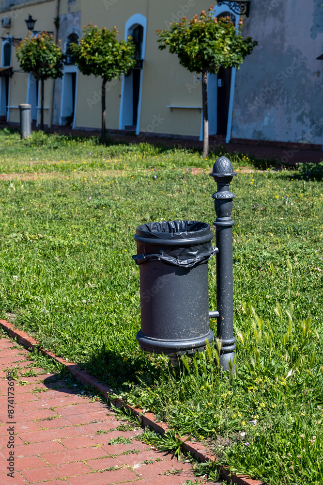 Obraz premium Public rubbish bin in a park - Serbia. Empty trash can in the park. Black bin in park. Black metal garbage bin in park.