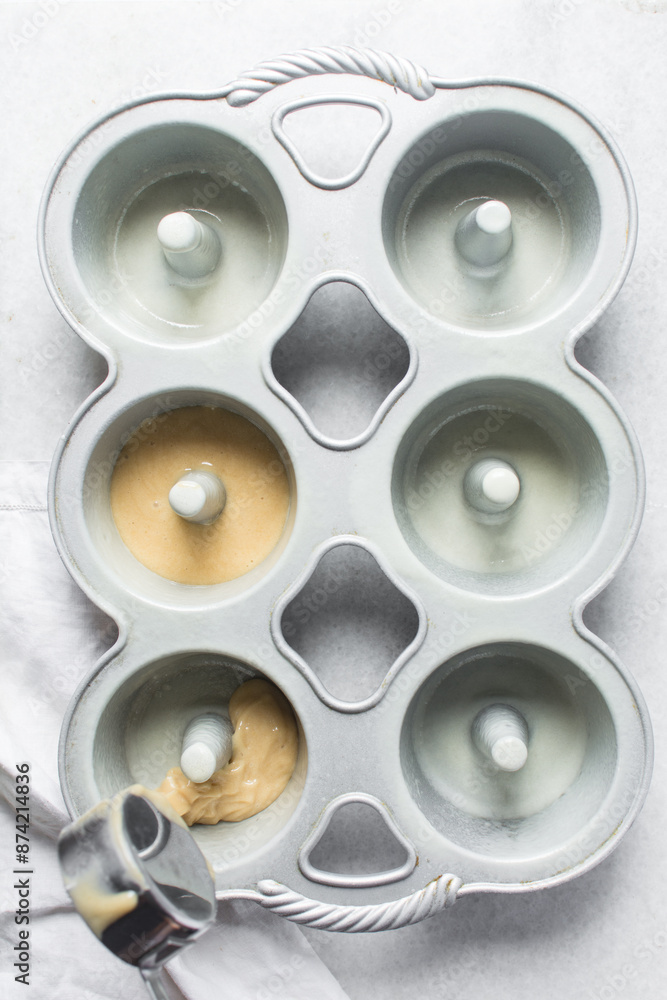 Putting cake batter in a mini bundt pan, Overhead view of vanilla cake batter in mini bundt cake pan, top view of cake batter in silver bundtlette baking pans, process of making mini bundt cakes Putting cake batter in a mini bundt pan, Overhead view of vanilla cake batter in mini bundt cake pan, top view of cake batter in silver bundtlette baking pans, process of making mini bundt cakes