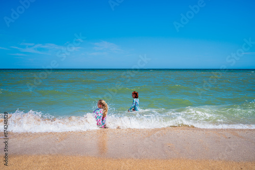 two children, a red-haired girl and a blonde girl on the seashore play in the waves and run along the beach on the sand, sit in the water in nature