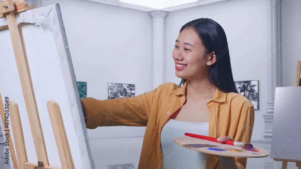 Asian Female Artist Showing Thumbs Up Gesture And Smiling While Painting On Canvas By Oil Paints And Brush In Gallery Asian Female Artist Showing Thumbs Up Gesture And Smiling While Painting On Canvas By Oil Paints And Brush In Gallery