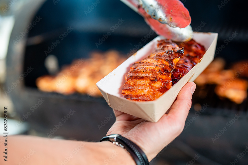 Φωτογραφία Close-up of a hand holding grilled chicken skewers in a paper tray, with a blurred barbecue grill in the background