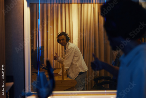 Young singer standing in recording room behind the glass and showing thumbs up to composer