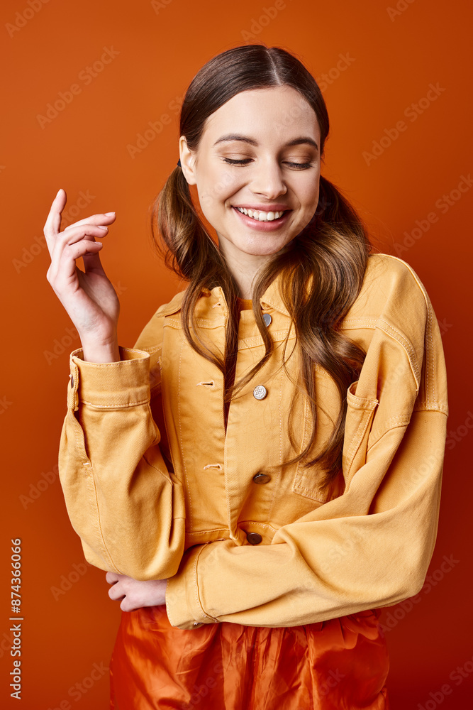 A stylish young woman in her 20s wearing a yellow shirt and pants poses in a studio with an orange background. A stylish young woman in her 20s wearing a yellow shirt and pants poses in a studio with an orange background.