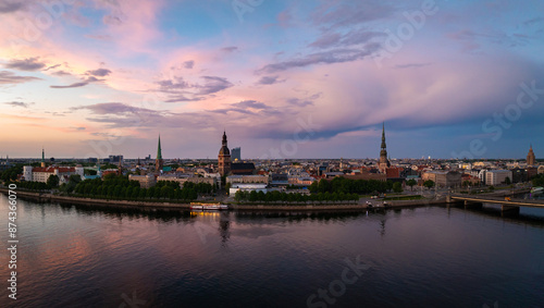 A stunning aerial view of Riga, Latvia, showcasing the city skyline with St. Peter's Church and Riga Dome Cathedral at sunset. The sky is painted in pink and purple hues.