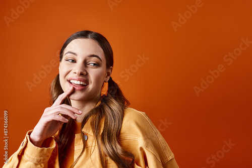Billede på lærred A young, stylish woman in her 20s strikes a pose with her finger on her lips against an orange studio backdrop