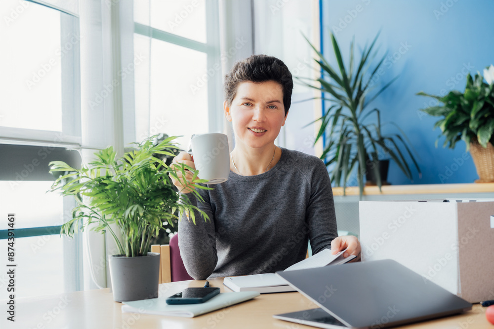 Satisfied smiling newcomer middle aged neutral gender female employee holds coffee cup and sitting at her new office desk work space. First working day concept. Dreaming about new opportunities Satisfied smiling newcomer middle aged neutral gender female employee holds coffee cup and sitting at her new office desk work space. First working day concept. Dreaming about new opportunities