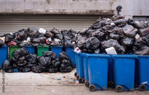 Photography A pile of overflowing black garbage bags spilling over the top of several blue and green recycling bins on a city street