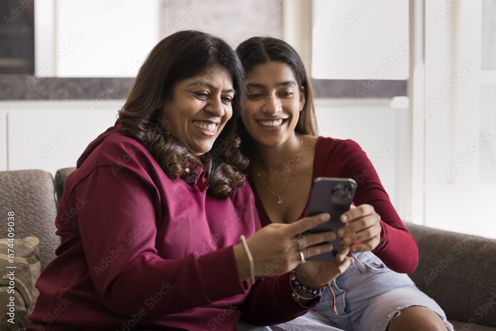 Happy young Indian daughter woman showing application on smartphone to cheerful mature mom. Mother and adult kid using mobile phone for Internet communication, laughing, having fun Happy young Indian daughter woman showing application on smartphone to cheerful mature mom. Mother and adult kid using mobile phone for Internet communication, laughing, having fun