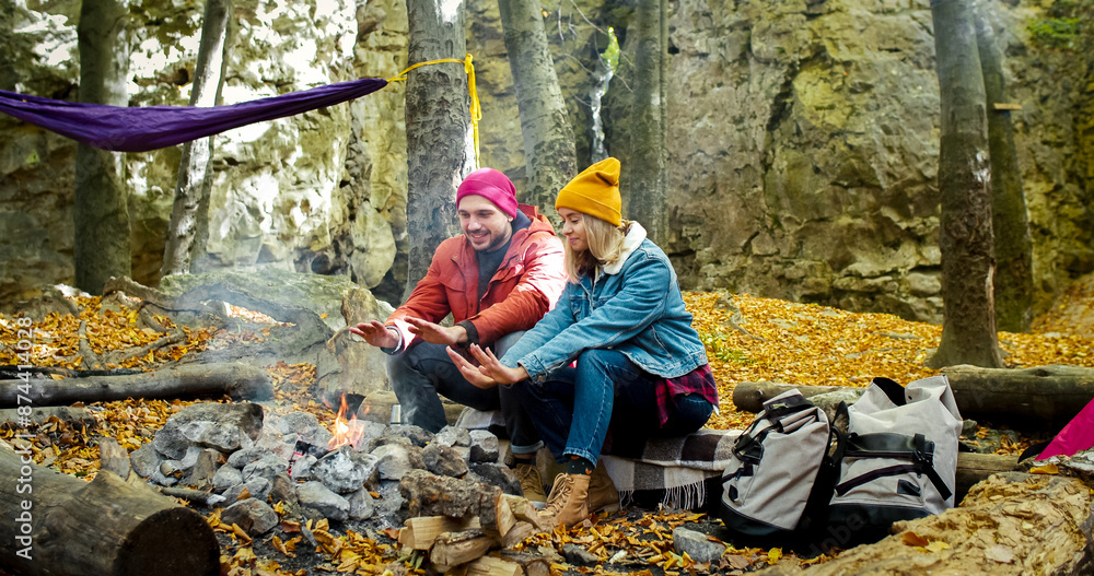 Couple of Caucasians sit near bonfire in forest. Man and woman warm hands by fire in the middle of autumn. On the background hammock hangs between trees. Couple of Caucasians sit near bonfire in forest. Man and woman warm hands by fire in the middle of autumn. On the background hammock hangs between trees.