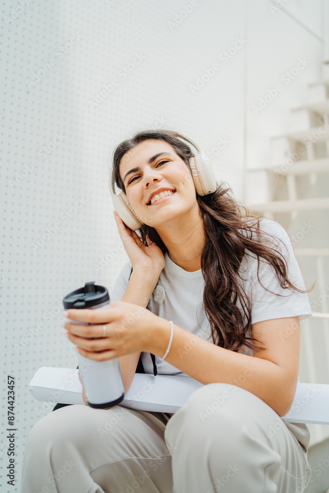 One young caucasian woman student sitting on stairs near university listening to music and drinking coffee One young caucasian woman student sitting on stairs near university listening to music and drinking coffee