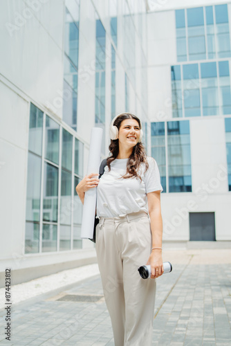 One young caucasian woman student going to university while listening to music on wireless headphones and holding papers and coffee 