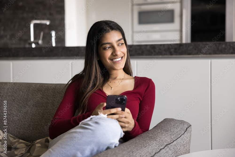 Happy pensive beautiful Indian woman holding smartphone, relaxing on comfortable home couch, looking away with toothy smile, using online app for communication, thinking Happy pensive beautiful Indian woman holding smartphone, relaxing on comfortable home couch, looking away with toothy smile, using online app for communication, thinking