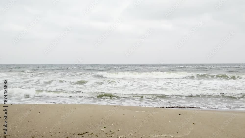 Stormy sea waves crashing on sandy beach with cloudy grey sky, showcasing coastal panorama in slowmotion Stormy sea waves crashing on sandy beach with cloudy grey sky, showcasing coastal panorama in slowmotion