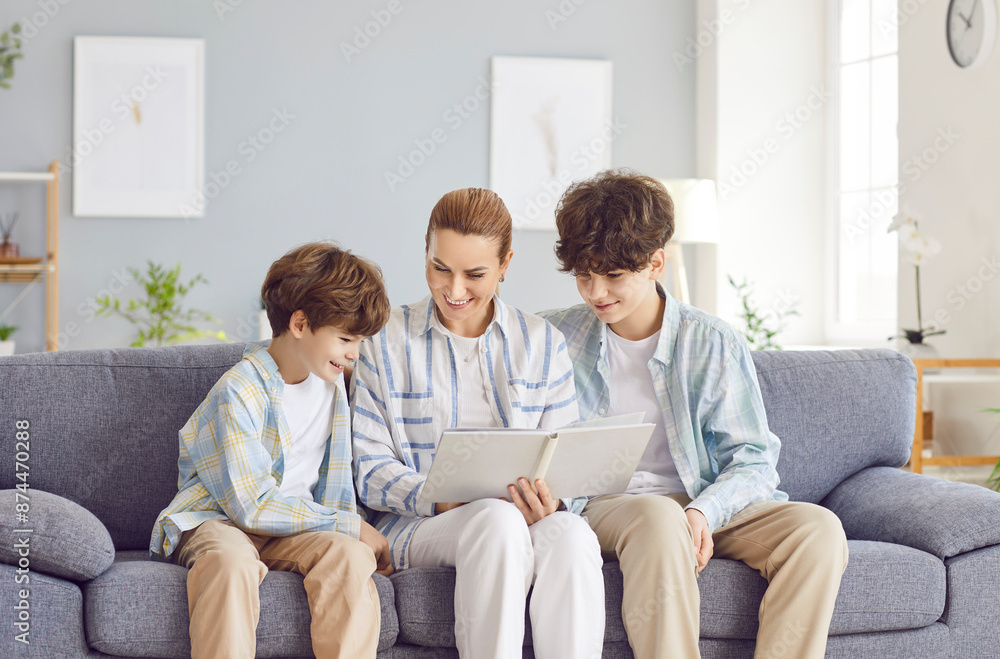 Portrait of a happy smiling young mother parent looking through a family photo album sitting on sofa at home with her children sons. Kids enjoying leisure time with mom. Family good memories