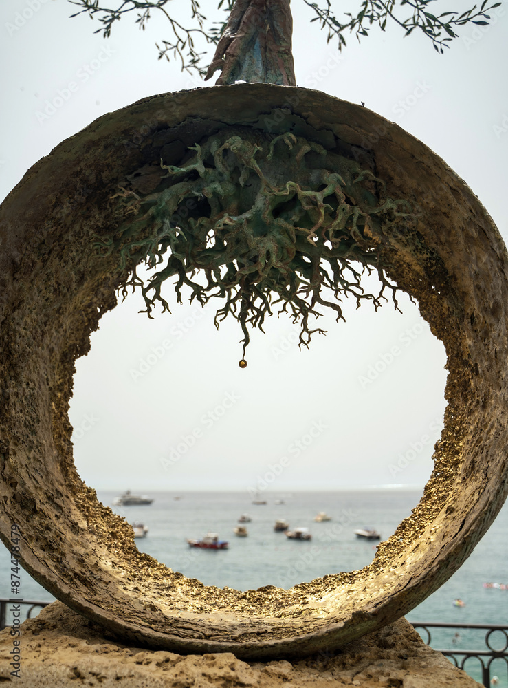 View of bunch of boats floating on a sea through a metal round circle with tree roots located in Amalfi town, Italy, Street art monument, Koi no Yokan aka premonition of love overlooking Amalfi Sea